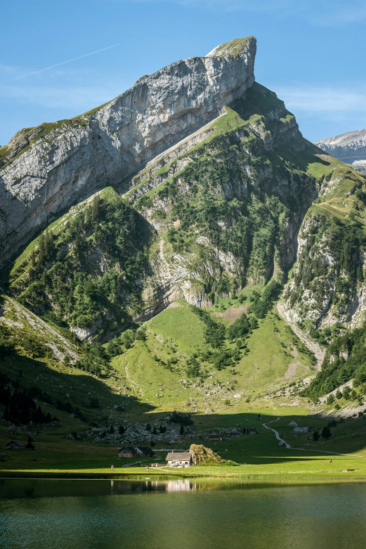 An alpine lake beneath a steep rocky mountain and green meadows